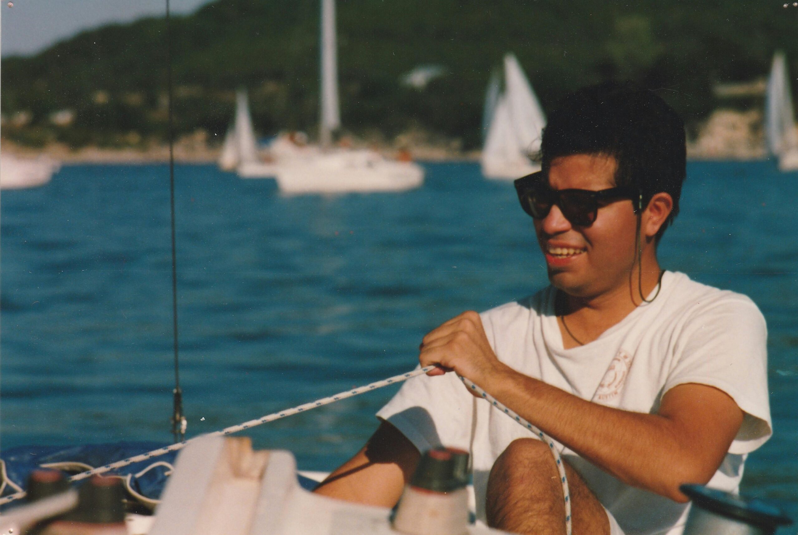 A man smiling while sailing on a sunny day with sailboats in the background.