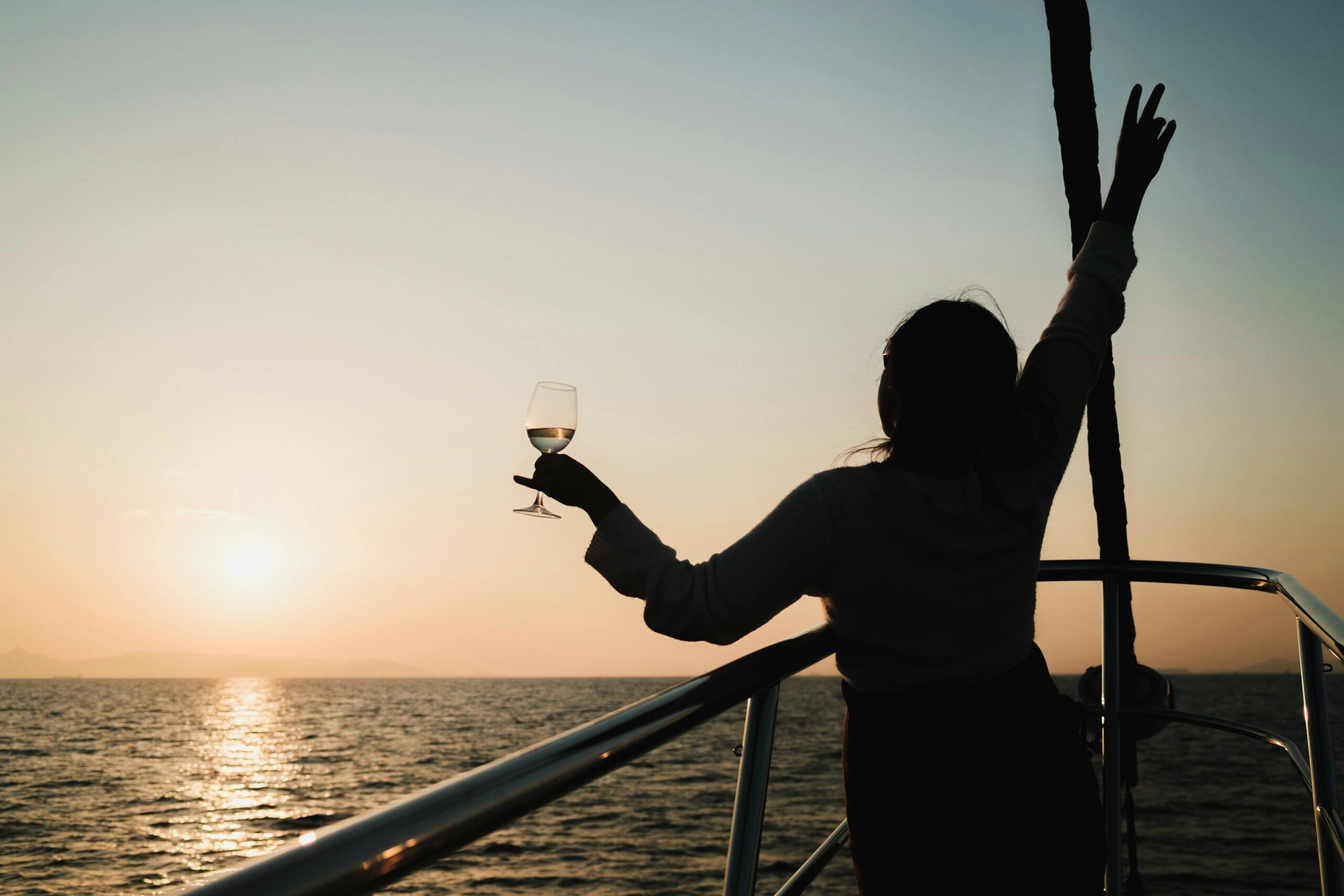Silhouette of a woman with a drink, celebrating a sunset on a yacht trip.